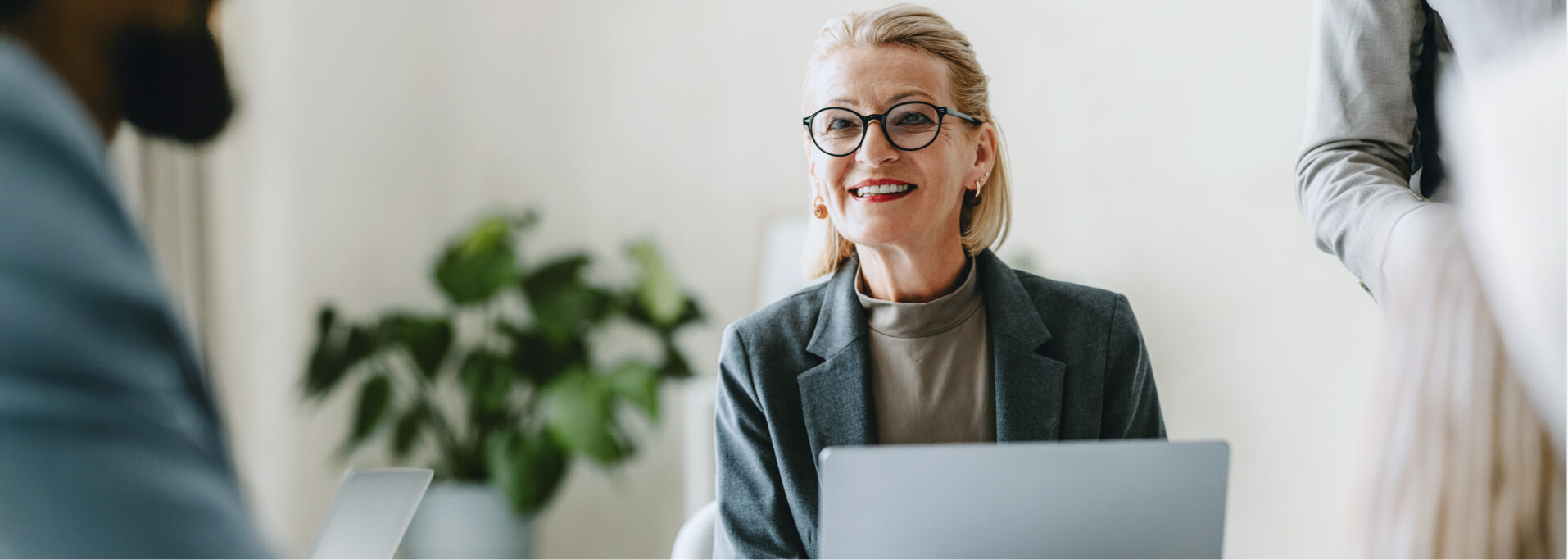 Lächelnde Frau mit Brille in einem Business-Outfit sitzt vor einem Laptop und spricht mit einem Kunden, der ihr gegenübersitzt.